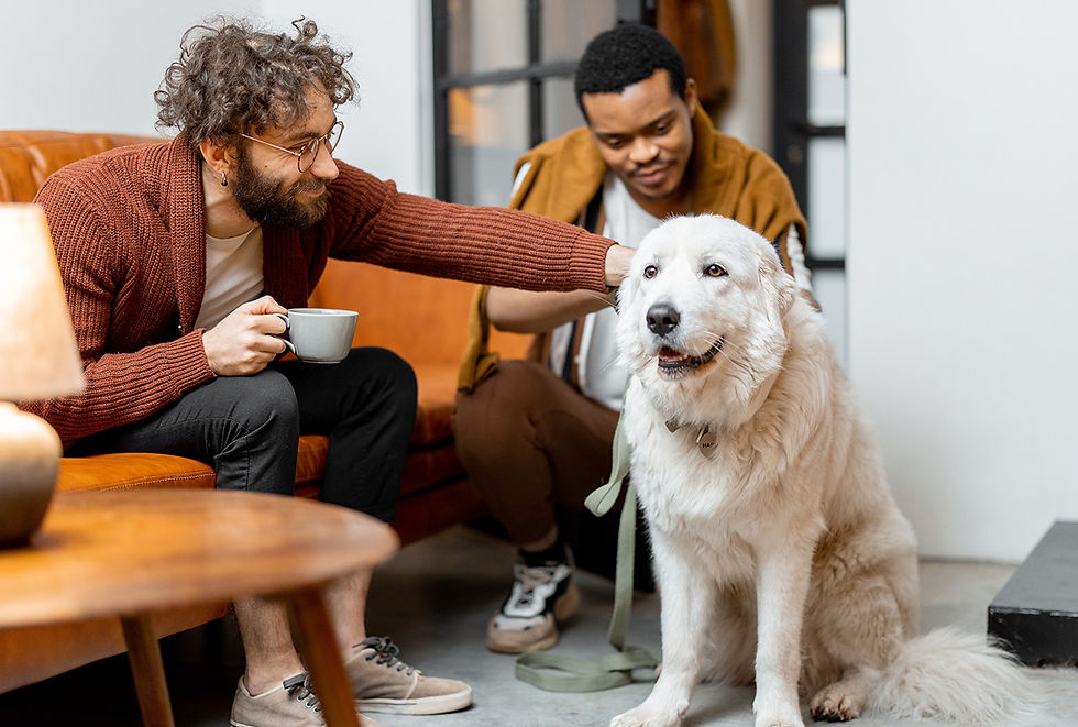 A man sitting on a couch, holding a coffee cup, smiles while petting a large white dog, while another man crouches down to pet the dog. 