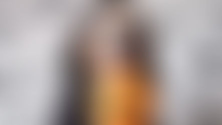 A dog in a black cape holds a pumpkin in their mouth.