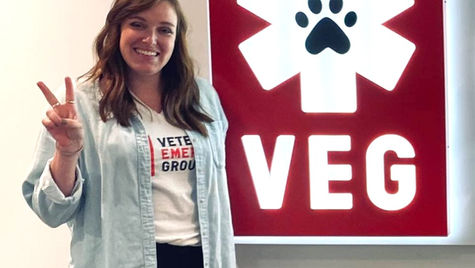 A smiling woman wearing a VEG t-shirt with a button down shirt over it, holding up two fingers next to a VEG sign