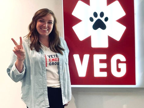 A smiling woman wearing a VEG t-shirt with a button down shirt over it, holding up two fingers next to a VEG sign
