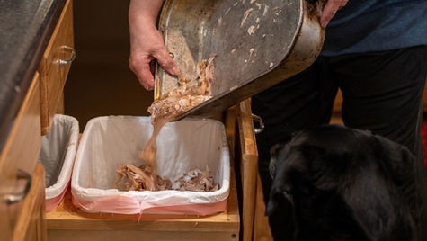 A person pours turkey pan contents into a garbage as dog watches