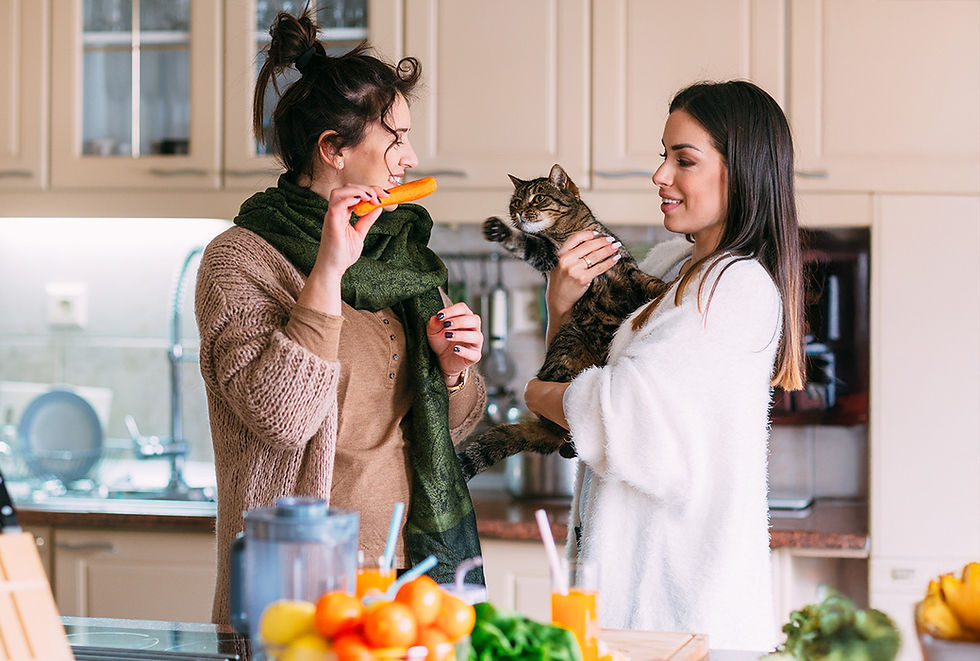 Two smiling women stand in a kitchen; one holds a tabby cat and the other holds a carrot, teasing the cat.