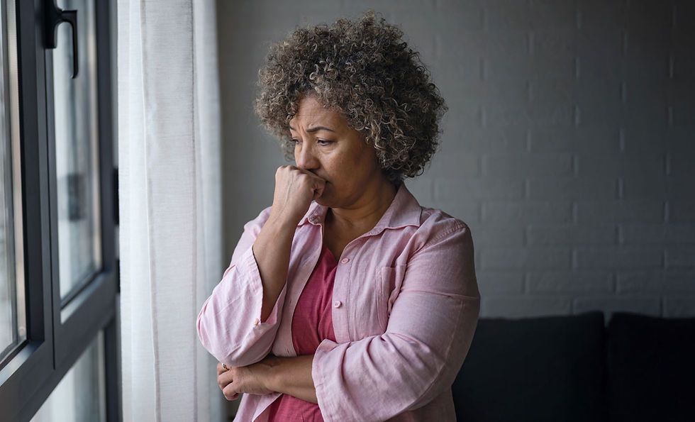 A women with curly hair and a pink shirt standing near a window with a sad look on her face.