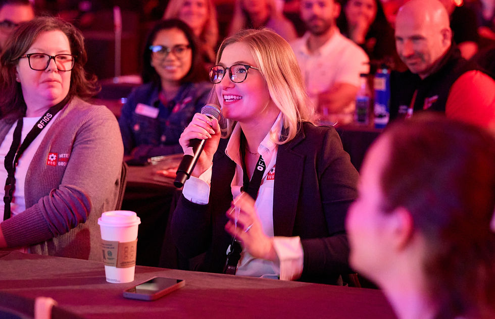 A woman with glasses and a microphone speaks at a table. Others listen, smiling.